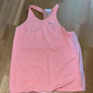 Rosa/ orange träningstopp från Nike - Säljer en laxrosa träningstopp från Nike med brottarrygg och ventilerande mesh på baksidan. Perfekt för gymmet eller löpning. Snygg och sportig design med Nike-logga framtill. storlek L barn men är som en XS.