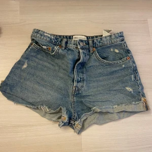 Blå jeansshorts från Zara - Säljer ett par blå jeansshorts från Zara med råa, fransiga kanter och slitna detaljer. Shortsen har fem fickor och stängs med knapp . Perfekta för sommaren 💕