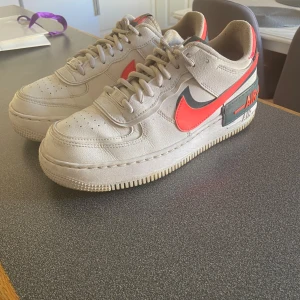 Nike Air Force 1 sneakers med röd swoosh - Säljer ett par Nike Air Force 1 sneakers i vit med röd och svart swoosh. Skorna har klassisk snörning, perforerad tå och tydlig AIR-detalj på sulan. Perfekta för dig som gillar streetwear och vill sticka ut med färg.