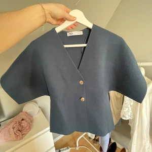 Mörkblå kortärmad blus från Zara - Säljer en stilren mörkblå blus från Zara med v-ringning och två dekorativa knappar framtill. Blusen har korta, vida ärmar och är tillverkad i ett mjukt, strukturerat material. Perfekt för dig som gillar minimalistisk stil.