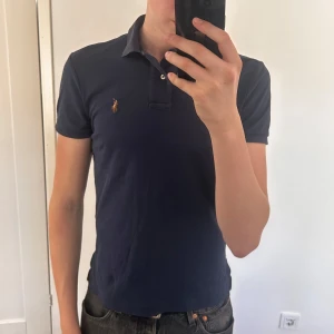 Mörkblå pikétröja från Ralph Lauren - Säljer en klassisk mörkblå pikétröja från Ralph Lauren med liten broderad logga på bröstet. Tröjan har korta ärmar, krage och knappar vid halsen. Perfekt för en stilren och avslappnad look. Använd men bra skick!