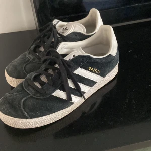Adidas Gazelle sneakers i svart och vitt - Säljer ett par klassiska Adidas Gazelle sneakers i svart mocka med vita detaljer och de ikoniska tre ränderna på sidorna. Skorna har snörning och platt sula, perfekt för en stilren vardagslook.