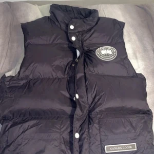 Svart dunväst från Canada Goose - Säljer en svart dunväst från Canada Goose med hög krage och vita tryckknappar. Västen har logotyp på bröstet och en patch med varumärket på fickan. Perfekt för kyliga dagar och enkel att matcha med olika outfits.