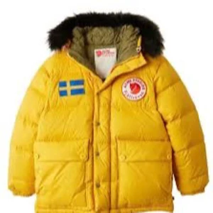 Gul dunjacka från Fjällräven - Gul dunjacka från Fjällräven med svart fuskpäls på huvan och svenska flaggan på bröstet. Jackan har tryckknappar, dragkedja och två stora fickor framtill. Perfekt för kalla dagar.