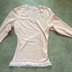 Ljusrosa långärmad topp med spetsdetaljer - Säljer en ljusrosa långärmad topp med v-ringning och fina spetsdetaljer vid halsringningen och nederkanten. Mjuk och följsam, perfekt för en söt och feminin look.