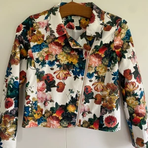 Blommig ungdomsjacka från Monsoon, 12-13 år - Färgstark jacka från Monsoon med blommigt mönster i rött, gult, blått och grönt på vit botten. Jackan har dragkedja framtill och klassisk krage. Perfekt för dig som vill sticka ut med en unik stil. Jackan är i storlek 12-13 år. Rökfritt hem.
