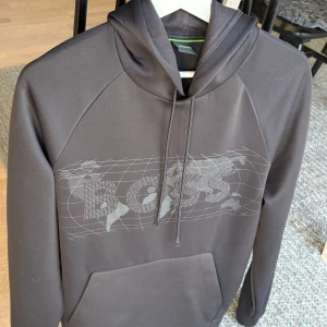 BOSS hoodie - Svart hoodie från BOSS med stort grafiskt tryck över bröstet och klassisk känguruficka. Tröjan har huva med dragsko och är tillverkad i ett mjukt syntetmaterial. Perfekt för en avslappnad och stilren look. Köptes för 2200 kroner på Johnells, 