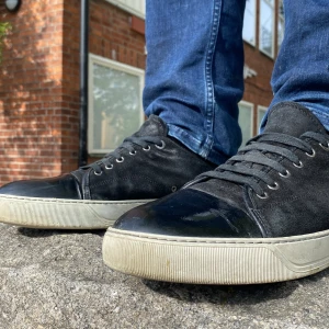 Svarta sneakers från Lanvin med mockadetaljer - Säljer ett par svarta sneakers från Lanvin med ovandel i mocka och detaljer i blankt skinn framtill och baktill. Skorna har vita, platta sulor och klassisk snörning. Perfekta för dig som gillar stilrena och tidlösa sneakers.