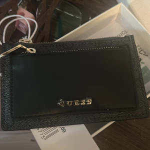 Svart plånbok från Guess - Säljer en stilren svart plånbok från Guess med guldfärgad logga och dragkedja. Plånboken har en slät framsida med Guess-detalj och mönstrad baksida med diskret logomönster. Alla klisterlappar , prislapp på, låda medföljer. Skulle säga att det mer är för mynt än kort men kort får absolut plats