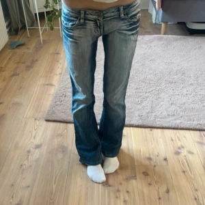 Blå bootcut jeans med slitningar - Säljer ett par blå bootcut jeans med snygga slitningar och ljusare partier framtill och baktill. Jeansen har låg midja, klassiska fickor och en lätt utsvängd passform nertill. Perfekta för en avslappnad och trendig look.