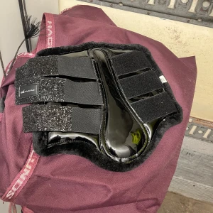 Svarta benskydd  - Säljer ett par svarta benskyddsett både bak och fram, med glittriga kardborreband och mjuk insida i fuskpäls. Skydden har en blank yta och är perfekt för tävling eller bara träning. Kommer i original genomskinlig väska. Aldrig använda. 