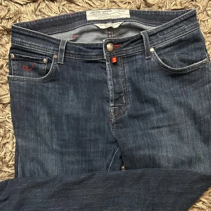 Mörkblå jeansbyxor från Jacob Cohen - Snygga mörkblå jeansbyxor från Jacob Cohen med klassisk femficksdesign och röda broderade detaljer. Byxorna har normal passform och stängs med knapp och dragkedja. Perfekta för dig som gillar stilrena jeans med unika detaljer.