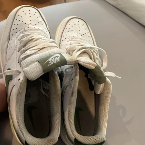 Nike Court sneakers i vitt och grönt - Säljer ett par fräscha Nike Court sneakers i vitt skinn med gröna detaljer och sula. Klassisk design med snörning, rund tå och grönt Nike-märke på hälen. Perfekta för dig som gillar stilrena och sportiga skor.