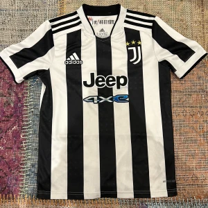 Juventus hemmatröja 2021/22 - Juventus officiella hemmatröja från säsongen 2021/22 med klassiska svartvita ränder, Adidas-logga, Jeep-sponsor och tre stjärnor ovanför klubbmärket. Tröjan har korta ärmar och är tillverkad i ett lätt och ventilerande material.