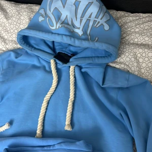 Blå SYNA tracksuit  - Snygg ljusblå tracksuit från SYNA med vit broderad text och snöre. Hoodie-1500 byxor-1200 ,båda-2500
