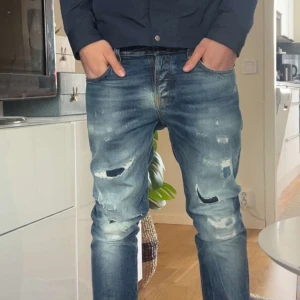 Blå slitna jeans från Nudie Jeans - Säljer ett par blå jeansbyxor från Nudie Jeans med slitningar och hål på framsidan. Byxorna har raka/slim ben, orangea sömmar. Perfekta för en avslappnad och trendig look. Nypris ca 2500kr