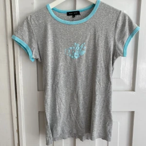 Grå t-shirt med turkosa detaljer från Bik Bok - Grå t-shirt från Bik Bok med turkosa kanter vid halsringning och ärmslut. T-shirten har ett litet turkost surfer tryck framtill 🩵den är lite sliten därav små hål i kragen.