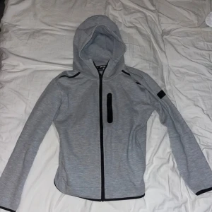 Ljusgrå hoodiejacka från Drytex - Säljer en ljusgrå hoodie från Drytex som är köpt på stadium. Hoodien har en hel ragkedja och svart detaljerad bröstficka med dragkedja. Hoodien har huva och är långärmad. Hoodien passar perfekt och är perfekt till både träning och vardag. Hoodien har använts ca 2-3 gånger. Storleken på Hoodien är Medium (M)