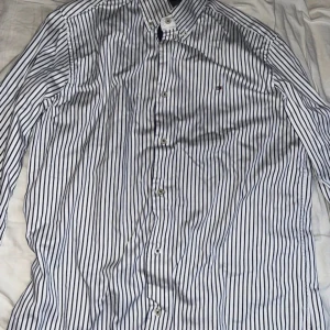  Tommy Hilfiger skjorta  - Säljer en vit skjorta med  marinblå ränder från Tommy Hilfiger. Skjortan har en krage med knappar på och en liten Tommy Hilfiger logga broderad på bröstet. Skjortan är väldigt skön att ha på sig och har använts ca 2-3 gånger. Storleken på Skjortan är Medium (M)
