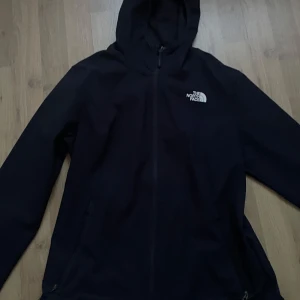 Svart vindjacka med huva från The North Face - Säljer en svart vindjacka från The North Face med huva och dragkedja framtill. Jackan har logga på bröstet och ryggen, samt två sidofickor med dragkedja. Perfekt för blåsiga dagar och enkel att matcha med olika outfits.