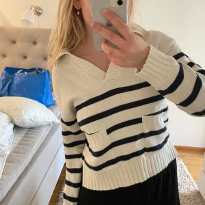 Randig stickad tröja från Holly & Whyte - Säljer en krämvit stickad tröja från Holly & Whyte med marinblå horisontella ränder. Tröjan har v-ringning, två fickor framtill och långa ärmar. Perfekt för en avslappnad och stilren look.