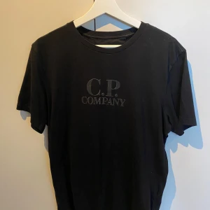 Svart t-shirt från C.P. Company - Svart t-shirt från C.P. Company med klassisk rund hals och tryckt logga på bröstet. Enkel och stilren design, perfekt för dig som gillar streetwear. Tillverkad i mjuk bomull och har korta ärmar.