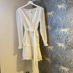 🌟Vit omlottklänning från Dennis Maglic 🌟 - Superfin vit kort omlottklänning från Dennis Maglic med långa ärmar och volangdetaljer. Klänningen har knyt i midjan och är gjord i ett glittrigt, lätt material som ger en lyxig känsla. Perfekt för sommarens alla tillfällen! (aldrig använd av mig, men köpt på vinted)