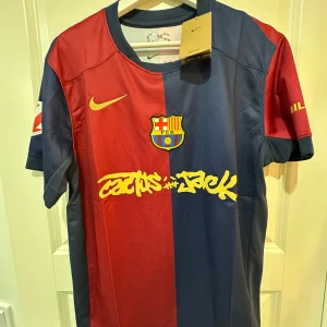FC Barcelona x Cactus Jack Nike tröja - Snygg FC Barcelona fotbollströja från Nike i samarbete med Cactus Jack. Tröjan har klassiska blå och röda paneler, gul Nike-logga och klubbmärke på bröstet samt Cactus Jack-tryck i gult över magen. Tillverkad i lätt och ventilerande material, perfekt för match eller streetwear.