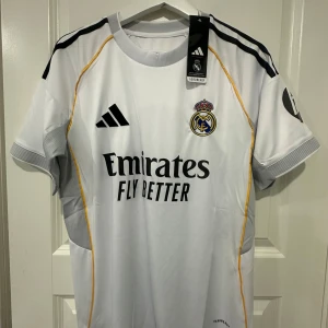 Real Madrid vit matchtröja Adidas 25-26 - Snygg vit Real Madrid fotbollströja från Adidas med svarta detaljer och gula linjer längs sidorna. Tröjan har klubbmärke på bröstet, Adidas-logga och sponsortryck. Tillverkad i lätt och ventilerande material, perfekt för träning eller match.