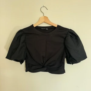 Svart croppad topp Barrels and Oil - Svart croppad topp från Barrels and Oil med puffiga korta ärmar och twistad detalj framtill. T-shirten har rund halsringning.