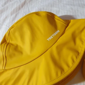 Gul regnhatt från Tretorn 48/50 - Snygg gul regnhatt från Tretorn i storlek 48/50. Hatten har bred brätte, tryckknappar och grått mjukt foder på insidan. Perfekt för regniga dagar och skyddar bra mot väta. Klassisk Tretorn-logga på utsidan.