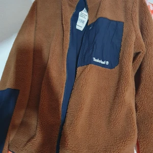 Brun teddyjacka Timberland S - Snygg brun teddyjacka från Timberland i storlek S. Jackan har marinblå detaljer på bröstficka och ärmar, dragkedja framtill och hög krage. Perfekt för kyliga dagar och ger en cool, avslappnad vibe. Ficka med dragkedja på bröstet och broderad Timberland-logga.