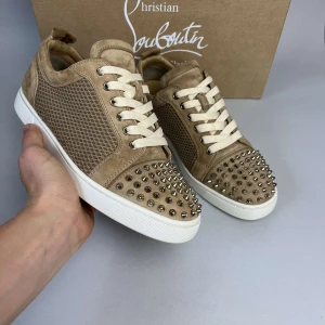 Beige Christian Louboutin sneakers med nitar - Snygga beige sneakers från Christian Louboutin i mocka med coola silvriga nitar på tån och klassisk röd sula. Skorna har vita snören och ett diskret mönster på sidorna. Perfekta för dig som vill sticka ut med exklusiv streetstyle. Boxen fås inte med på grund av borttappad