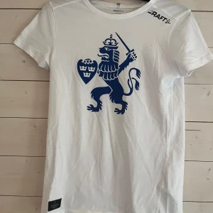 Vit t-shirt med blått lejon IFK Göteborg XS - Vit t-shirt IFK Göteborg i storlek XS med ett stort blått lejontryck på bröstet och diskret logga på axeln. Klassisk rund hals och korta ärmar. Tillverkad i mjuk bomull, perfekt för dig som gillar sportig och stilren design.