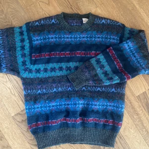 Mönstrad ulltröja i blått och rött - Retro stickad tröja i 100% ull med färgglada mönster i blått, rött, grönt och lila. Tröjan har rund hals, långa ärmar och ribbade muddar. Står att tröjan är large, men upplevs som mycket mindre! 