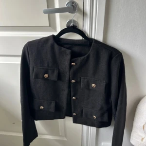 Svart kavaj med guldknappar - Stilren svart kavaj med fyra fickor framtill och snygga guldknappar. Croppad modell med rund halsringning och lång ärm. Perfekt för dig som gillar klassisk men trendig stil.