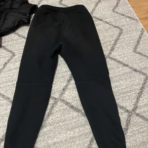 Svarta mjukisbyxor från Nike - Svarta mjukisbyxor från Nike med dragsko i midjan och diskret Nike-logga på ena benet. Byxorna har en smal passform, dragkedjeförsedda fickor och är tillverkade i en mjuk bomullsblandning. Perfekta för chill eller träning.