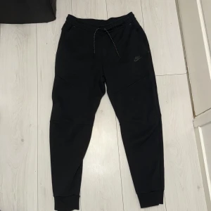 Svarta mjukisbyxor från Nike - Svarta mjukisbyxor från Nike med dragsko i midjan och ribbade muddar vid bensluten. Byxorna har en clean look med diskret Nike-logga på låret och är tillverkade i mjukt bomullsmaterial. Perfekta för chill eller träning.