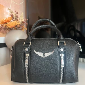Svart handväska med silverdetaljer - Snygg svart handväska i skinn med två rejäla handtag och silverfärgade dragkedjor framtill. Väskan har en cool vingdetalj i silver på framsidan som ger en unik touch. Perfekt storlek för det viktigaste och passar till många olika outfits.