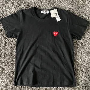 Svart Comme des Garçons Play t-shirt - Svart t-shirt från Comme des Garçons Play med det ikoniska röda hjärtat med ögon broderat på bröstet. Klassisk rund hals, korta ärmar och mjuk bomullskvalitet. Enkelt men ändå riktigt snygg statement-piece. Storlek L och helt oanvänd.