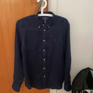 Mörkblå skjorta från Polo Ralph Lauren S - Klassisk mörkblå skjorta från Polo Ralph Lauren i storlek XL barn men sitter som en S. Skjortan har långa ärmar, knappar framtill och ikonisk röd broderad logga på bröstet. Tillverkad i bomull och passar perfekt till en stilren look.