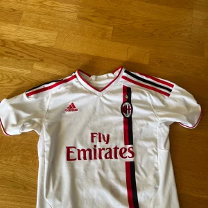 AC Milan Seedorf 10 bortatröja Adidas - Snygg vit retro AC Milan bortatröja från Adidas med röda och svarta detaljer, klubbmärke på bröstet och Fly Emirates-tryck. Ryggen har Seedorf och nummer 10 i rött. Klassisk V-ringning och korta ärmar, tillverkad i lätt och ventilerande material.