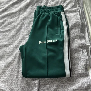 Gröna Palm Angels mjukisbyxor - Snygga gröna mjukisbyxor från Palm Angels med vit sidoremsa och svart detalj. Byxorna har elastisk midja och logga tryckt på ena benet. Perfekta för en chill och sportig look.