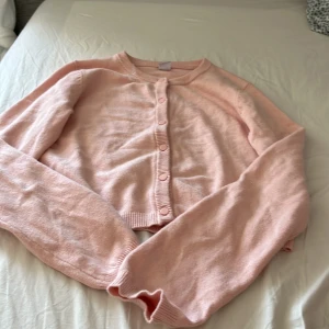 Ljusrosa kofta från Lindex, Kids 170 - Supersöt ljusrosa kofta från Lindex i storlek 170. Koftan är stickad, har rund halsringning och knäpps med matchande knappar framtill. Perfekt att slänga över en topp när det är lite kyligt ute.