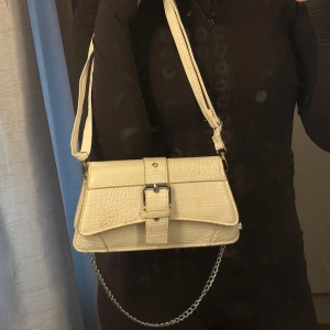 Beige axelväska med krokoprägling - Säljer en beige axelväska med krokodilpräglat mönster och snyggt spänne framtill. Väskan har justerbar axelrem och en cool silverfärgad kedjedetalj. Perfekt storlek för det viktigaste och ger en trendig touch till din outfit.