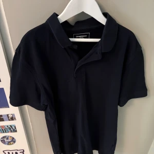 Mörkblå pikétröja från Dressmann - Snygg mörkblå pikétröja från Dressmann i klassisk modell med krage och korta ärmar. Perfekt för dig som gillar en stilren och enkel look. Tröjan är gjord i mjuk bomull som känns skön mot huden.