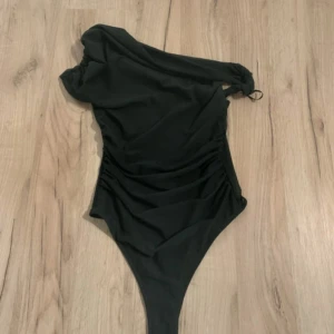Svart oneshoulder bodysuit - Snygg mörkgrön bodysuit med oneshoulder-design och draperade detaljer längs sidan. Toppen har en ärm och knyt vid axeln för extra stilpoäng. Perfekt för dig som vill sticka ut med en cool och modern look.