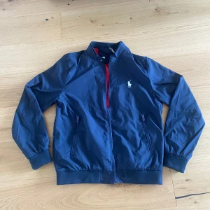 Ralph Lauren vindjacka - Säljer denna vind/regn jacka från Ralph Lauren i storlek XL men passar M. Jackan har aldrig använts och har kvar originaletiketterna. Väldigt stilren, speciellt till kyligare sommarkvällar samt även fram mot hösten.