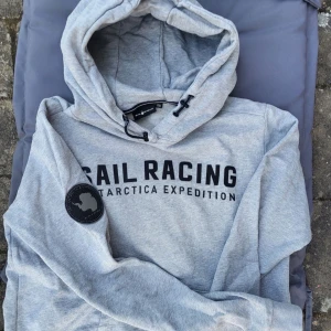Grå Sail Racing hoodie med tryck - Snygg ljusgrå hoodie från Sail Racing med svart tryck på bröstet och patch på ärmen. Tröjan har huva med snörning och är tillverkad i mjuk bomullsmix. Perfekt för dig som gillar sportig och avslappnad stil. Passar både xs och s då den är rätt stor i storlek.