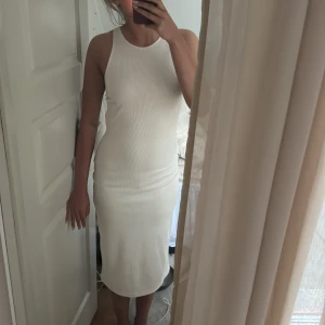Vit ribbad klänning med öppen rygg - Supersnygg vit ribbad klänning i midi-längd med tight passform. Klänningen är ärmlös och har en cool öppen rygg som ger en extra touch. Perfekt för dig som gillar stilrena och trendiga plagg. Materialet är stretchigt och följsamt.